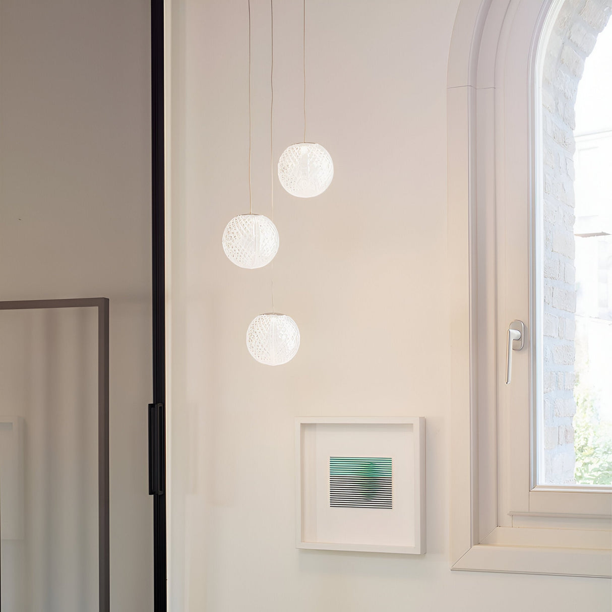A minimalistic interior showcases the Lustris 3 Light LED Cluster Pendant in Silver, featuring crystal-effect diffusers elegantly suspended from the ceiling, alongside a small framed abstract artwork on the wall and a window with an arched top that allows natural light to flow in for energy efficiency.