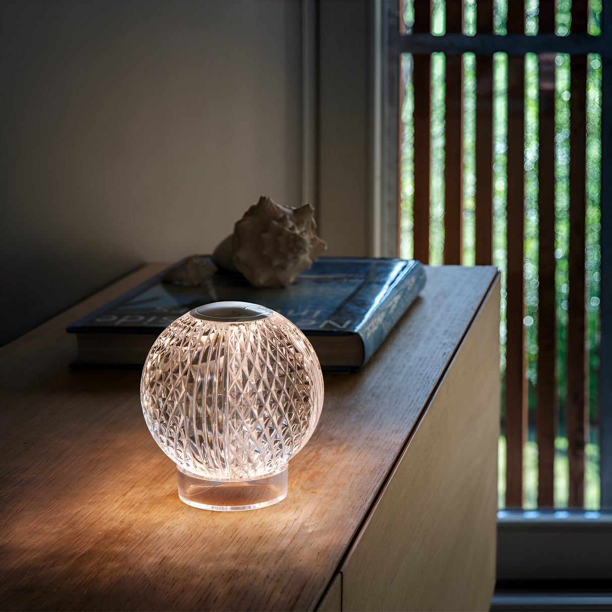 A glowing, spherical Lustris LED Table Lamp 2.2W 3000K - Clear with a textured surface sits on a wooden table next to two closed books and a seashell. This lamp, with its soft light filtering through vertical blinds, creates a cozy ambiance in the room.