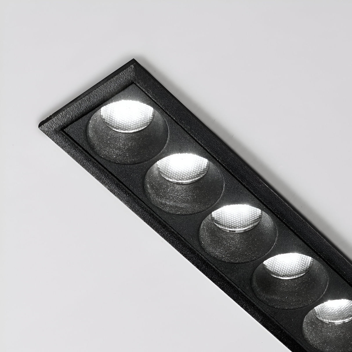The Phosix Slim LED Recessed Downlight, a sleek rectangular black fixture with five 10W, 3000K circular lights, is mounted on the ceiling. Its energy-efficient lighting creates a bright glow on the plain white surface, showcasing a modern and slim design.