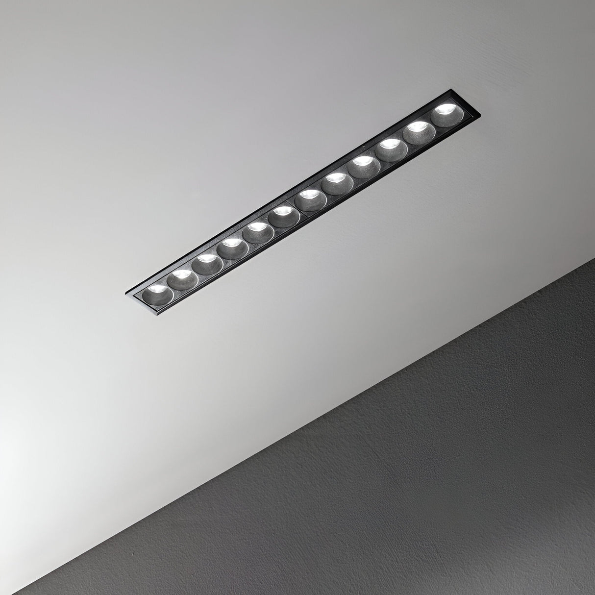 A contemporary, rectangular recessed light fixture integrated into a gray ceiling showcases the energy-saving Phosix Slim LED Recessed Downlight. With its sleek design, this 20W, 3000K black fixture features multiple round bulbs that emit a gentle glow, highlighting the ceiling's smooth, textured finish.