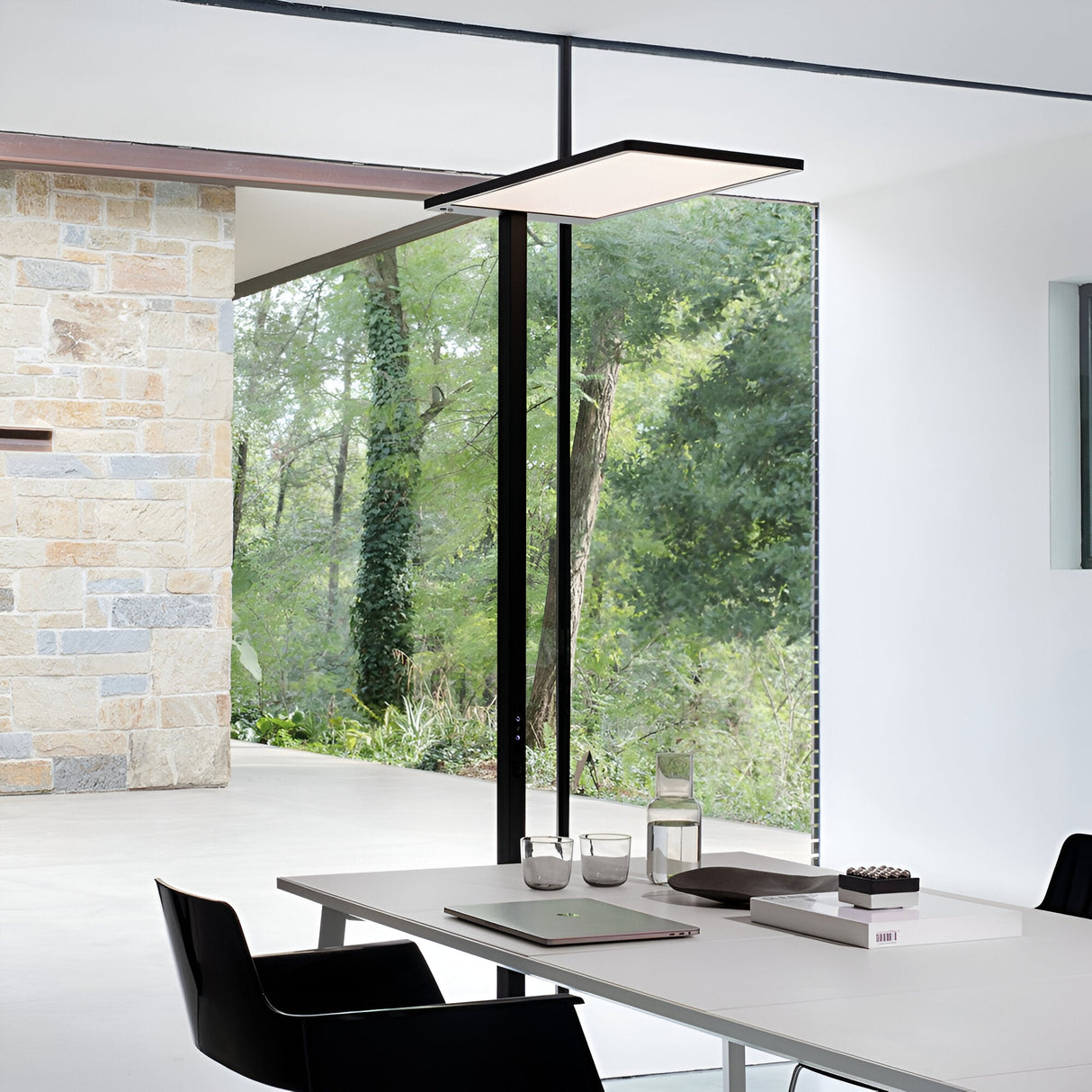 In a minimalist interior, a white table and black chairs complement the sleek overhead light fixture, creating an ideal setting for adjustable lighting. Large windows reveal a lush green forest outside. The modern decor features a stone wall detail on the left, enhanced by an elegant Luminor LED Floor Lamp 76W 4000K in black.