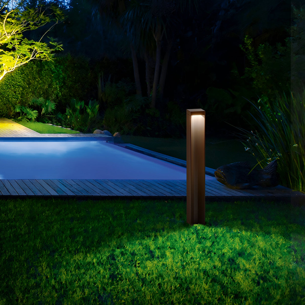 A serene night scene showcases a well-lit backyard with the Viritis Outdoor LED Bollard Light 12W 3000K in brown, crafted from powder-coated aluminium, illuminating a grassy area. A wooden deck encircles a softly illuminated swimming pool, surrounded by lush trees and bushes bathed in gentle ambient light.