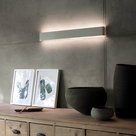 The Clarityx LED Wall Light 37W 4000K - White, 83cm brightens a concrete wall above a wooden sideboard with abstract art prints and dark ceramic vases and bowls, offering an energy-efficient and contemporary lighting solution.