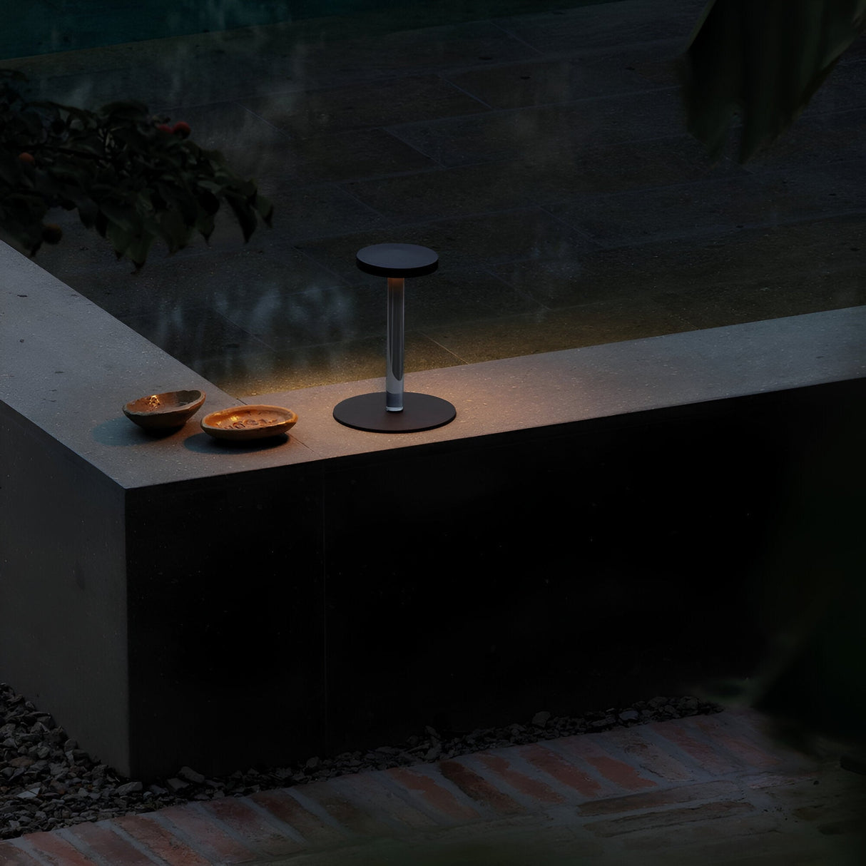 In a softly illuminated outdoor setting, the Solixis LED Table Lamp 3.6W 3000K in Black casts a warm glow on two shallow bowls resting on a stone ledge. Its minimalist design integrates perfectly with the serene backdrop of water and lush greenery, evoking a sense of tranquility during the evening.