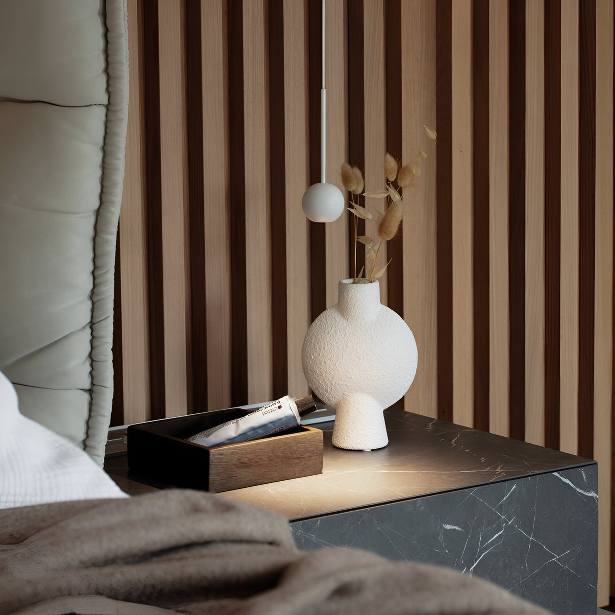 A modern bedroom nightstand featuring a black marble surface displays a white textured vase with dried flowers and a wooden tray holding various items. The Arclight LED 1 Light Sphere Pendant in white offers efficient lighting overhead, complemented by a wooden slat wall partially covered by a cushioned headboard, embodying modern interiors.