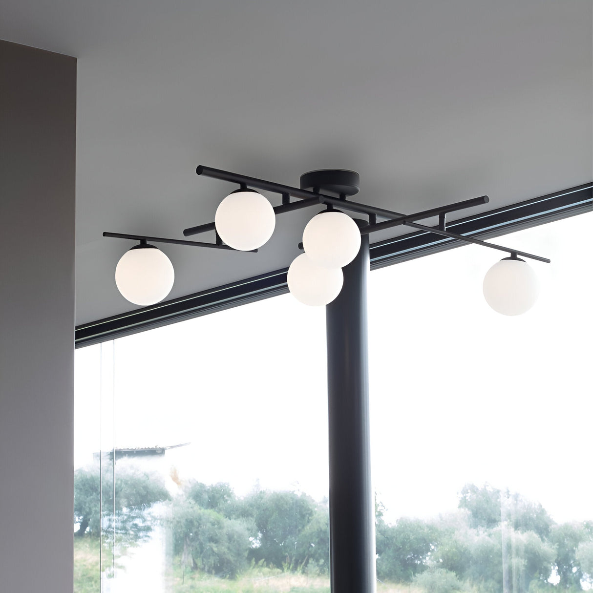 The Lucida 5 Light Semi Flush - Gold showcases a modern design with five spherical white bulbs and sophisticated white blown glass diffusers mounted on intersecting black metal rods. Ideally positioned near a large window, it enhances the room's minimalist decor while providing a view of trees outside.