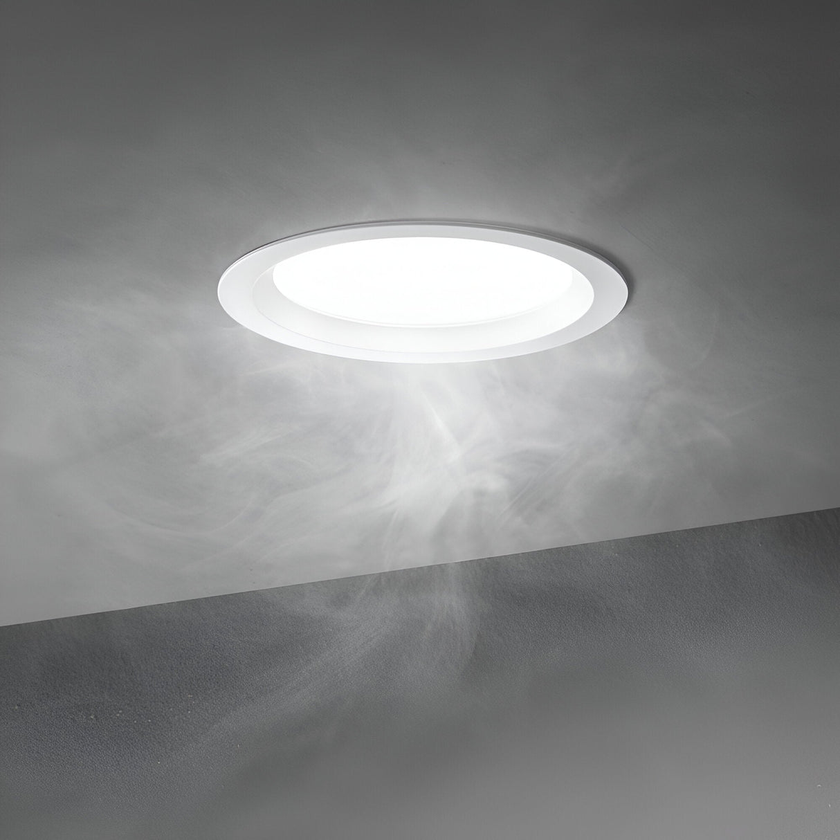 The Radiara-2 Round LED Recessed Downlight, 46mm-15W, emits a gentle, 3000K warm white light throughout the room. Delicate wisps of smoke or mist elegantly swirl around it, enhancing the serene and ethereal atmosphere against the smooth, gray ceiling.