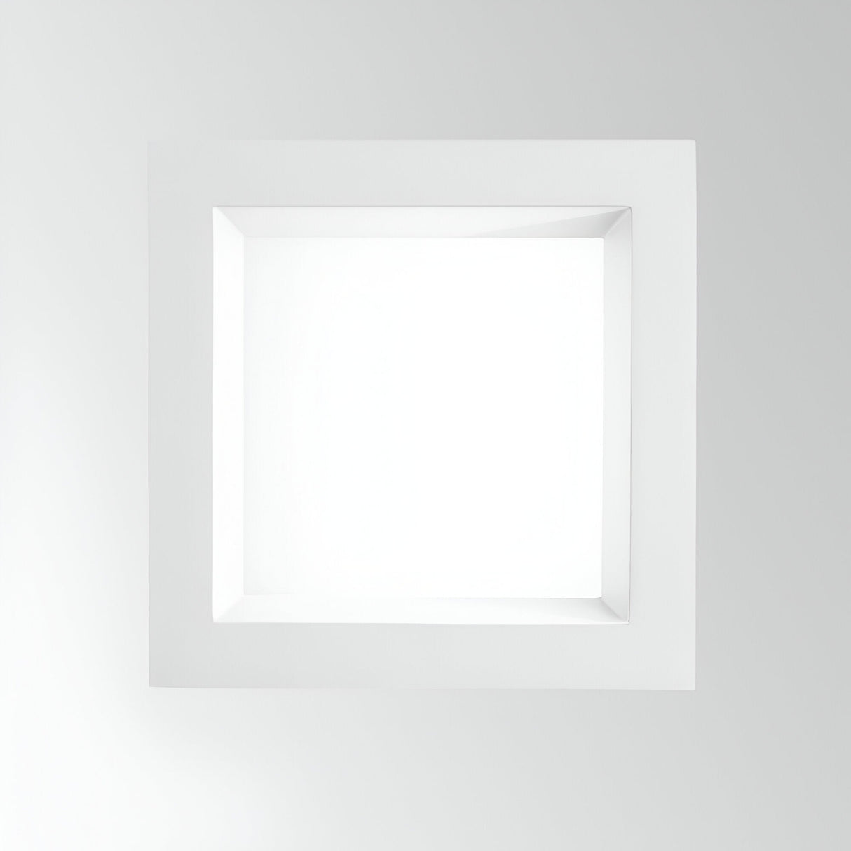 The Radiara-2 Square LED Recessed Downlight 167mm - 18W, 3000K, in white, serves as a minimalist architectural feature with its rectangular design and recessed, shadowed center set against a light gray background. Its clean lines and sharp angles offer depth and modernity, further enhanced by the soft glow from the IP65-rated LED bulb.