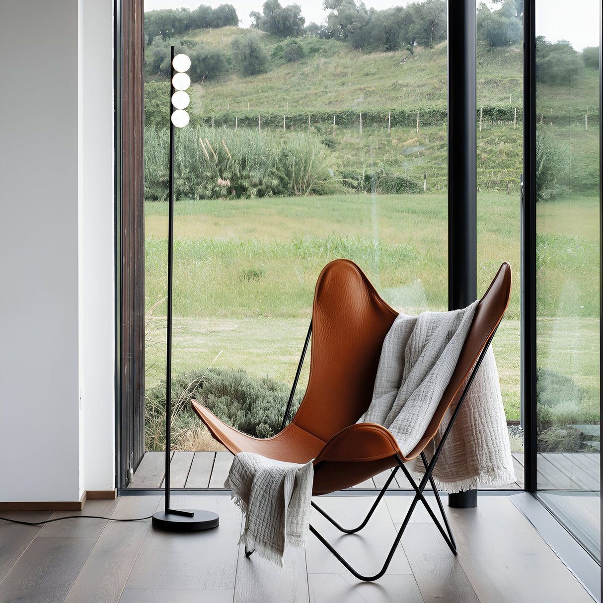 A modern living room exudes sophistication with a brown leather butterfly chair draped in a gray throw blanket. The Lyrica LED Floor Lamp 12W 3000K in black stands elegantly beside it, casting energy-efficient lighting. The room boasts large windows overlooking a green grassy field and hilly landscape.