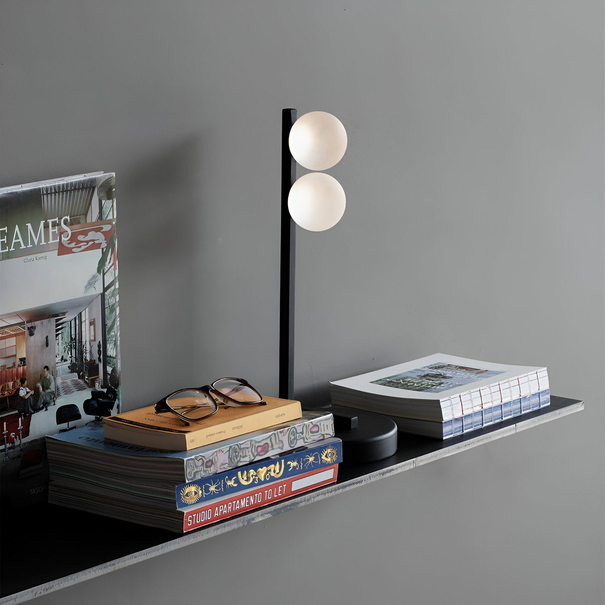 A modern wall shelf holds books, magazines, and a pair of eyeglasses. The Lyrica LED Table Lamp in black, featuring two round bulbs offering 6W energy-efficient lighting at 3000K, adds a sleek touch. The gray wall background enhances the contemporary elegance of the minimalist decor.