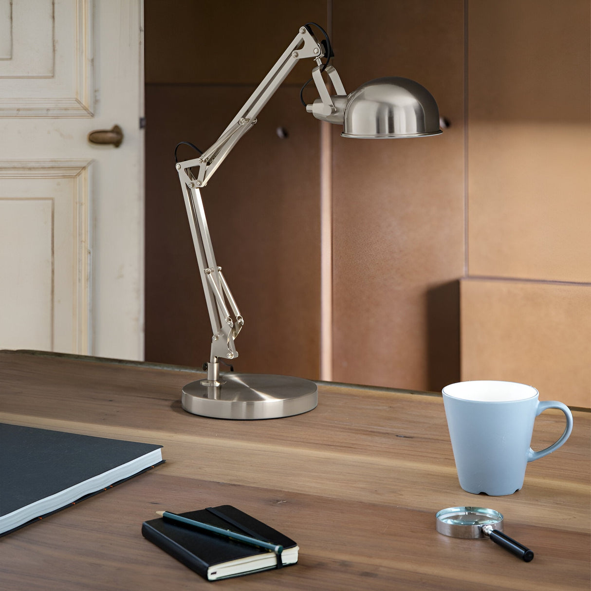 An elegant Orlix Table Lamp - Silver, featuring an adjustable arm, graces a wooden table. Beside the lamp, there's a closed black notebook, a magnifying glass, and a light blue cup. The backdrop includes a neutral-toned wall and the outline of a partially visible door.