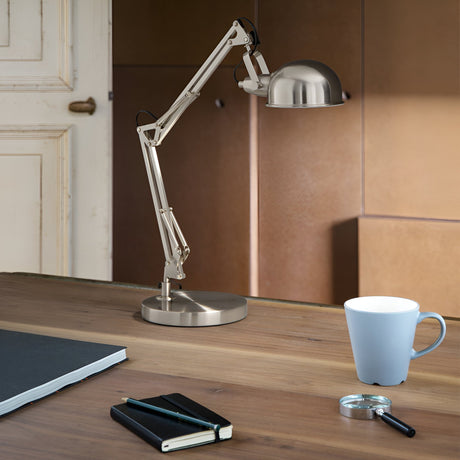 An elegant Orlix Table Lamp - Silver, featuring an adjustable arm, graces a wooden table. Beside the lamp, there's a closed black notebook, a magnifying glass, and a light blue cup. The backdrop includes a neutral-toned wall and the outline of a partially visible door.