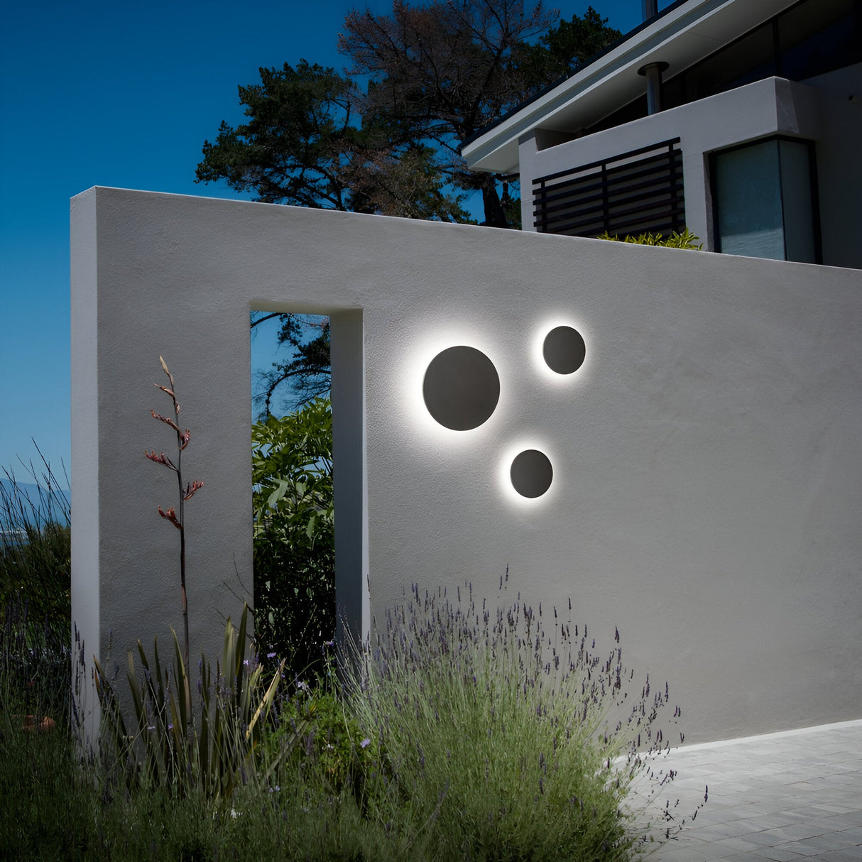 The Eclipseax Outdoor LED Wall Light 12W 3000K - Grey, standing at 24cm, casts a modern glow against the white surface with its sleek design and IP54 durability rating. Nearby, lush greenery and plants thrive, with tall trees in the background under a clear blue sky, enhancing the serene atmosphere.