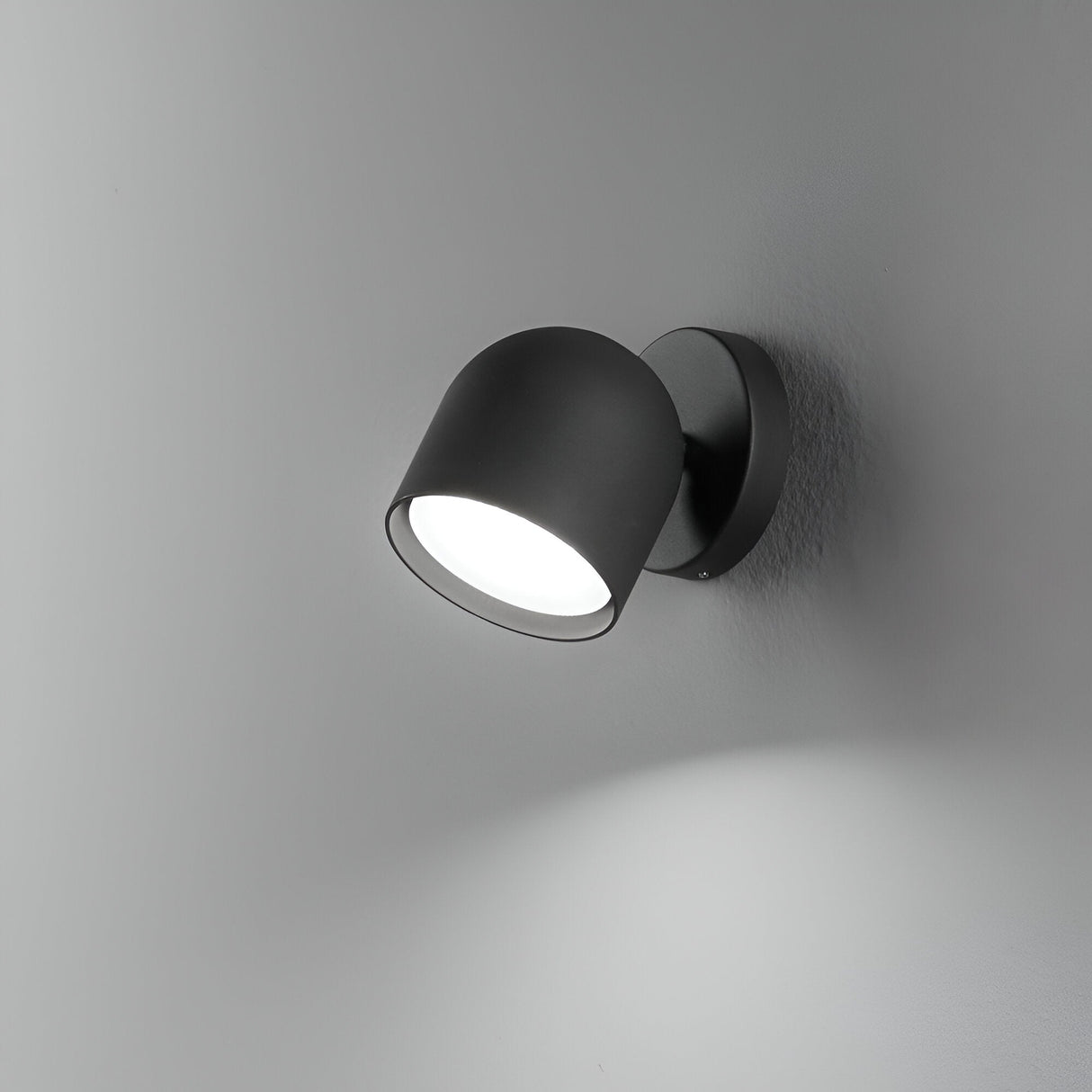 The Fable LED Single Spotlight in black, featuring a cylindrical shade and adjustable head, is mounted on a light gray wall. Its minimalistic design projects a circular beam of light downward, forming a soft shadow below.