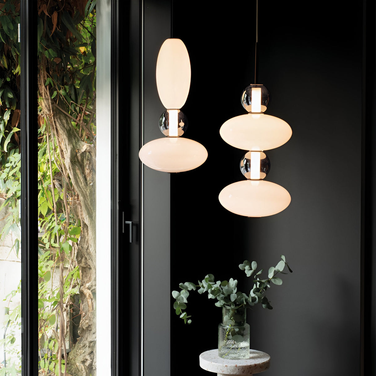 In a dimly lit room, two Gleara 1 Light LED Pendant lights with oval-shaped frosted glass hang gracefully. Beneath them, a small round table holds a glass vase with eucalyptus leaves, embodying minimalist design. A large window to the left presents the lush greenery outside, accentuated by energy-efficient lighting.