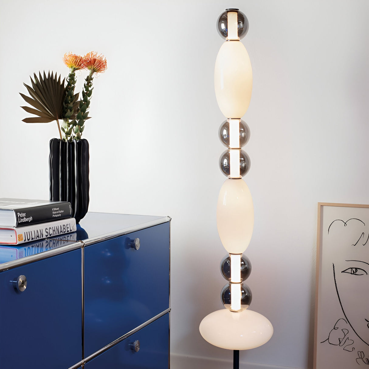 A modern room features the Gleara LED Floor Lamp in black and white, its tall, artistic design adding energy-efficient illumination with its oval shapes. A blue cabinet houses books and a black vase filled with flowers, while a minimalist line art portrait rests against the wall in this contemporary design space.