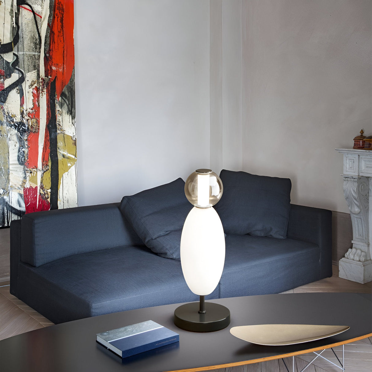 A modern living room showcases a sleek dark blue sofa, abstract red and white artwork, and a Gleara Table Lamp - White on the table. A decorative fireplace adds warmth, while a couple of books are neatly placed beside the table lamp.