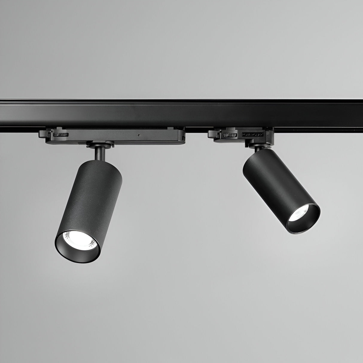 A pair of Glimmara LED Three Phase Track Spotlights, featuring a sleek black design and energy-efficient 21W power with a warm 3000K temperature, are mounted on a ceiling rail against a neutral gray background. The cylindrical heads are angled in different directions, allowing for adjustable illumination.