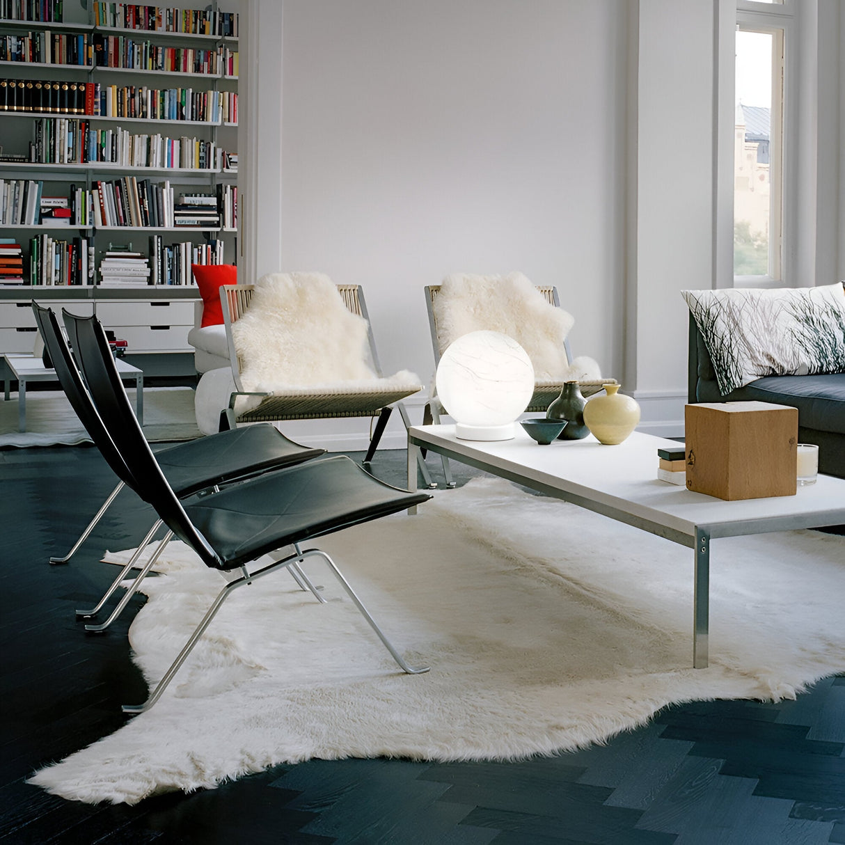 A stylish living room features contemporary interiors with modern black chairs, a white table adorned with a Gleonis Large Table Lamp - White, 20cm, and a sheepskin rug. Bookshelves filled with books line the wall. Decorative items accompany the lamp on the table as natural light streams through expansive windows.