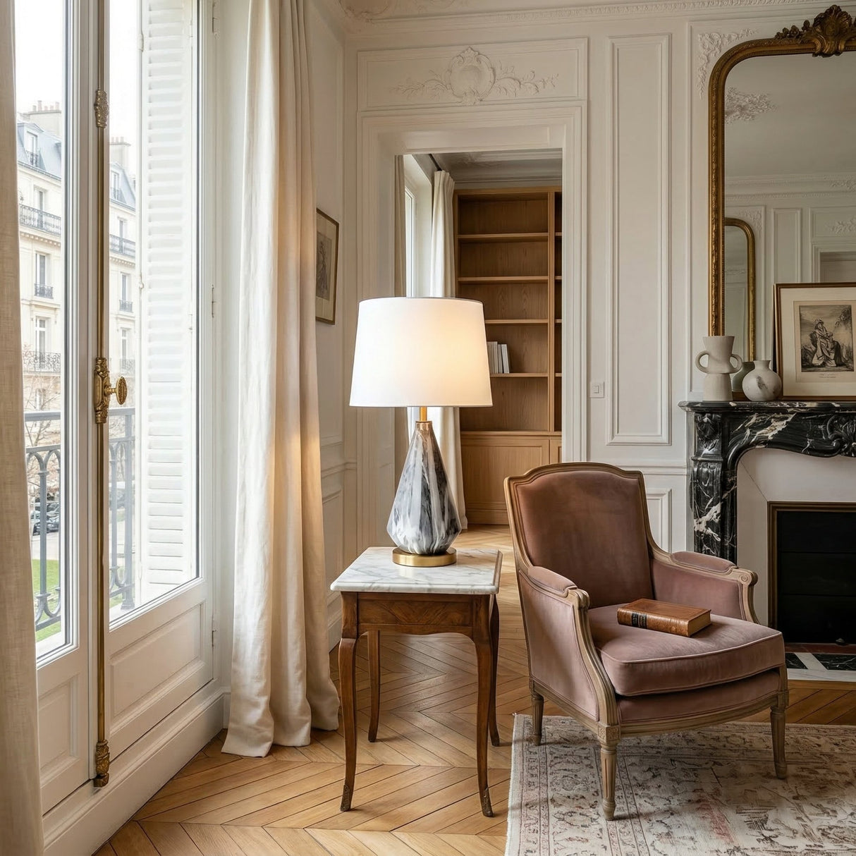 A cozy, elegant living room with an armchair, side table featuring the Calvero 1 Light Diamond Facet Marble Table Lamp in Black & Brass, marble fireplace, large mirror, bookshelves, and tall windows with white curtains letting in natural light.