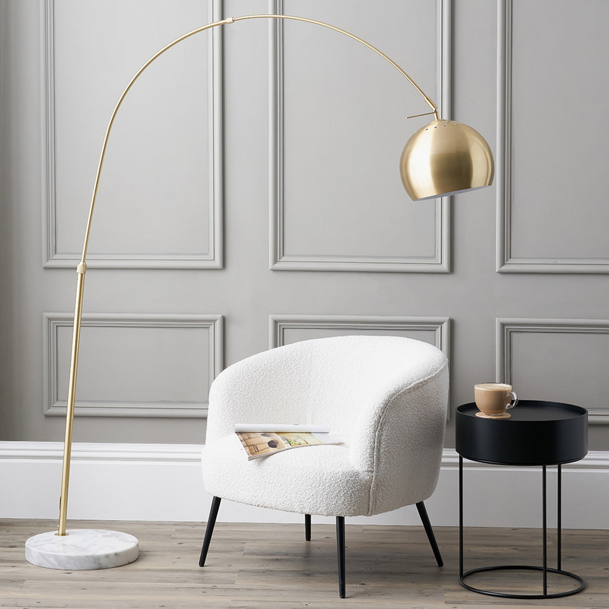 Feliciani brushed brass floor lamp with curved arm, round metal shade, and white marble base, shown beside a white boucle armchair and black side table.