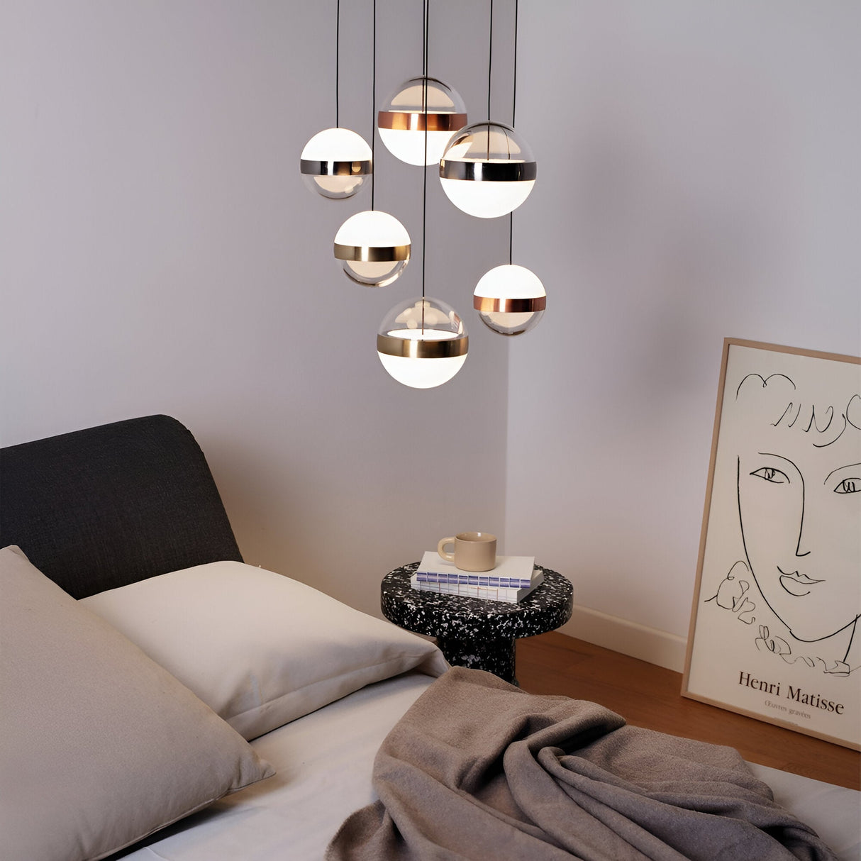 A modern living area features a black bed adorned with beige bedding and a snug brown blanket. A round black side table supports a coffee cup, while overhead, the Arionis 6 Light LED Cluster Pendant in white offers energy-efficient illumination at 58W and 3000K. A framed abstract line art is elegantly positioned on the floor.