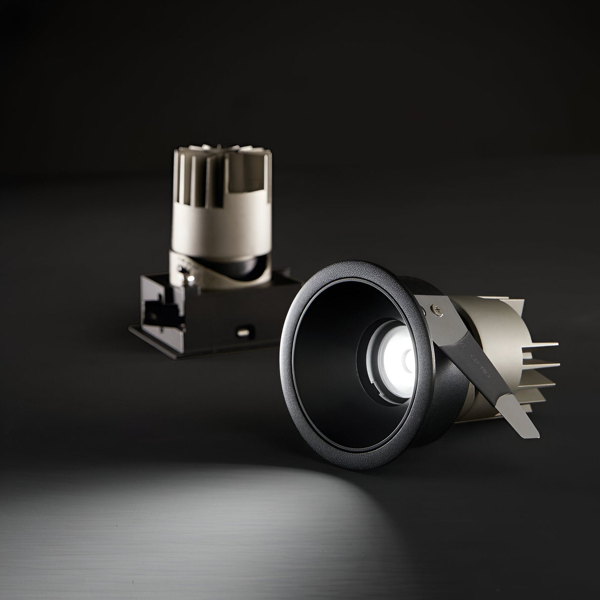 Two Glistenis Trim Square Frame 78mm recessed ceiling lights are depicted against a dark background. One, with a matte black finish, is positioned upright with its bulb glowing brightly. The other rests on its side, unlit. Both lights boast a modern design and sleek trim frame for stylish downlight installations.