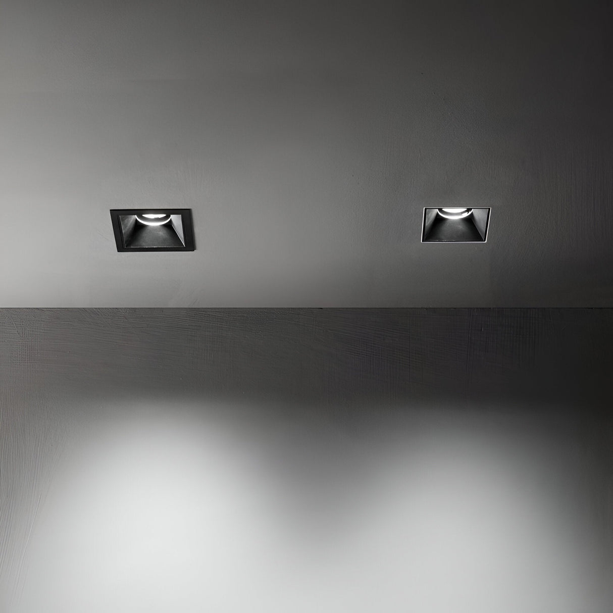 Two Glistenis Trim Square Frame 78mm recessed ceiling lights, featuring sleek black trim frames, are illuminated, casting soft light on a smooth, gray surface below. The scene is minimalistic, emphasizing the contrast between these downlight installations and the matte black finish of the dark surroundings.