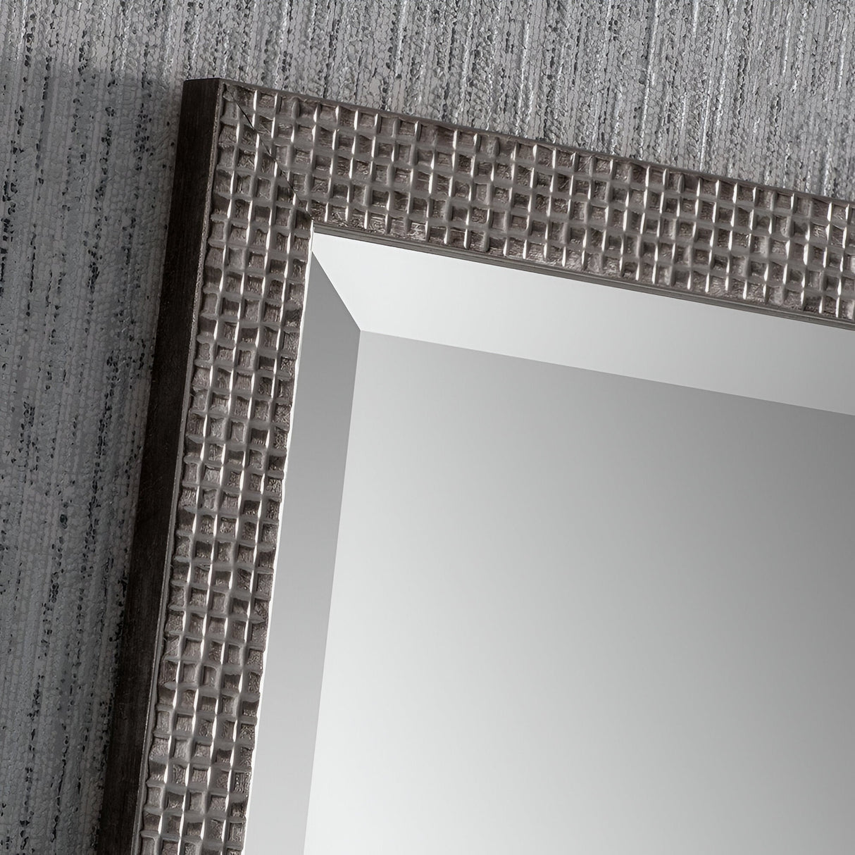 The Frost Rectangle Wall Mirror - Grey features a close-up of its textured frame with a checkered pattern, showcasing modern design. Angled against a gray, patterned wall, the mirror's reflective surface elegantly captures part of the wall's texture, making it a standout piece of home decor.