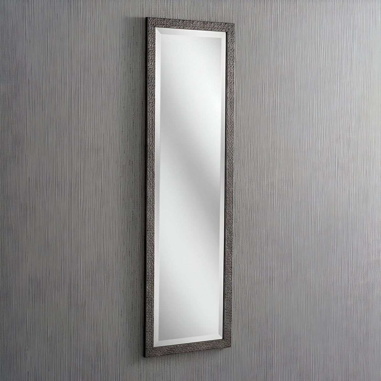 The Frost Rectangle Wall Mirror - Grey features a tall, rectangular design with a textured dark frame. Mounted vertically on a subtle light gray wall, it embodies modern design and reflects light to enhance brightness and space.