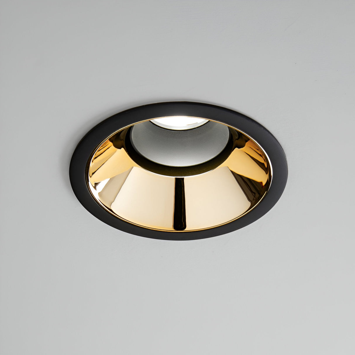 The Glistenis Trim Round Frame 109mm - Black showcases a recessed ceiling light with a glossy gold interior and a black finish rim, elegantly contrasted against a gray background. Perfect for sophisticated downlight installations.
