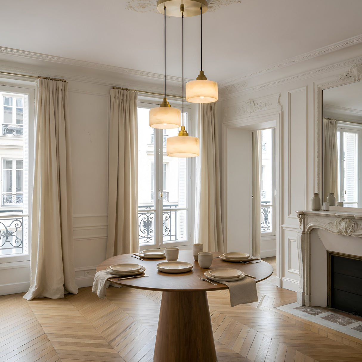A bright, elegant dining room with tall windows, beige curtains, a round table for four, a white fireplace, large mirror, and three Brinex 3 Light Cluster Alabaster Pendant Lights in soft white & brushed brass hanging above.