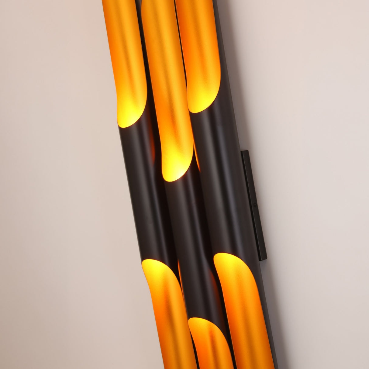 The Stealth 6 Light Wall Light in Brass & Black showcases three vertical black tubes emitting warm yellow-orange light through curved cutouts, creating a modern sculptural effect on any neutral wall.