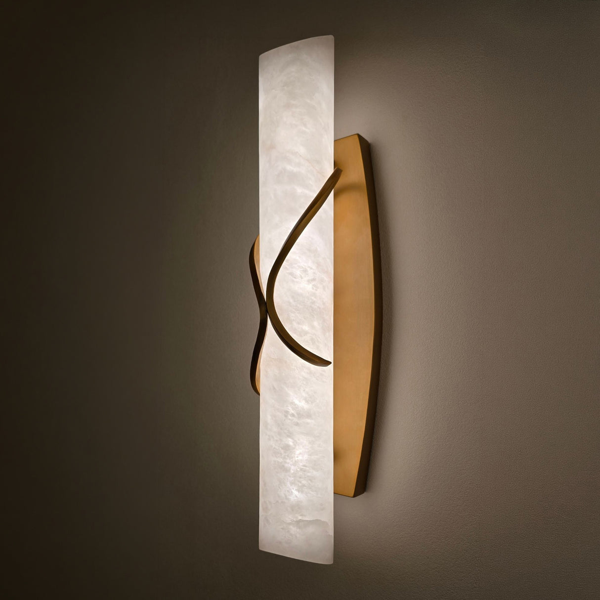 Valora LED wall light with a vertical alabaster column wrapped by curved brushed brass bands, illuminated warm white, mounted on a dark wall.
