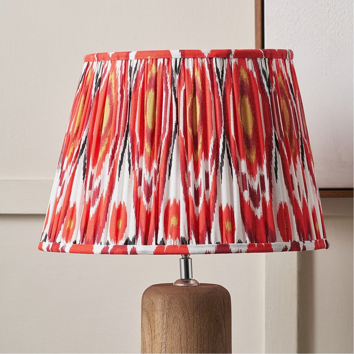 Izara Red Ikat Pleated Tapered Lampshade, 35cm, in red, black and yellow ikat pattern fabric, shown on a wooden lamp base against a neutral wall.