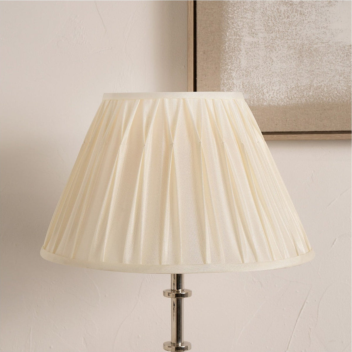 Garbo Cream Silk Mix Pinch Pleated Tapered Lampshade, 45cm, shown fitted on a chrome table lamp base against a plain wall with a framed artwork nearby.