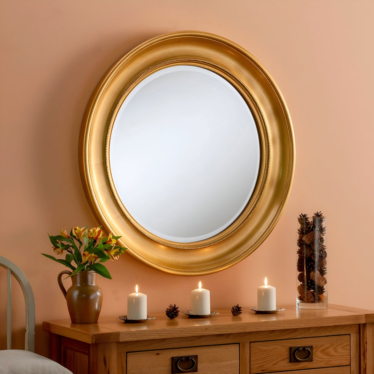 A Luxe Circle Wall Mirror Gold - 91cm with an elegant gold frame hangs on an orange wall above a wooden dresser. The dresser is adorned with a vase of flowers, three lit candles, and a decorative item.