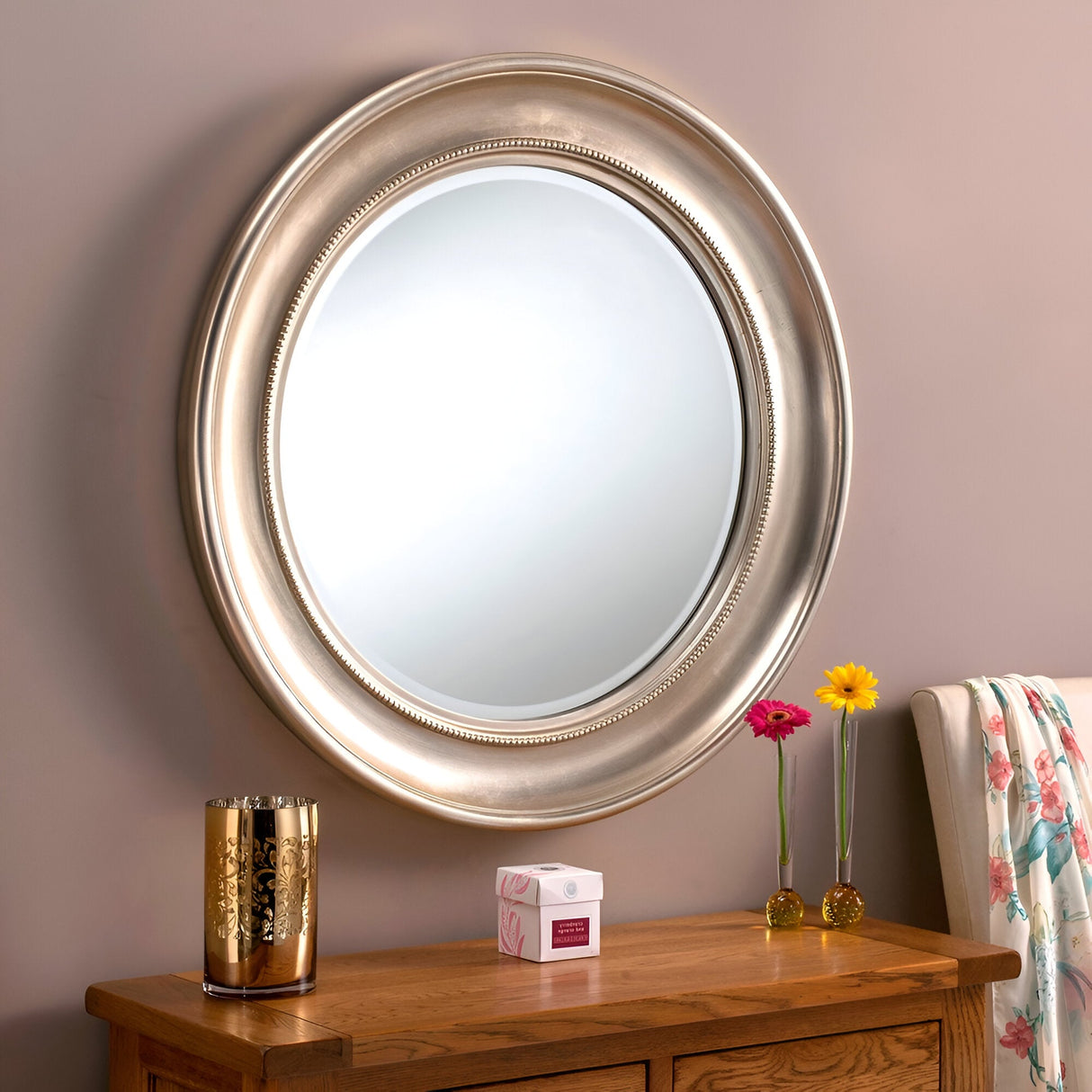 A Luxe Circle Wall Mirror Silver - 91cm adorns a beige wall above a wooden dresser. The dresser showcases a golden vase, a small white box, and two vibrant flowers in slender vases. A floral-patterned cloth is elegantly draped over the chair back, showcasing exquisite interior styles.