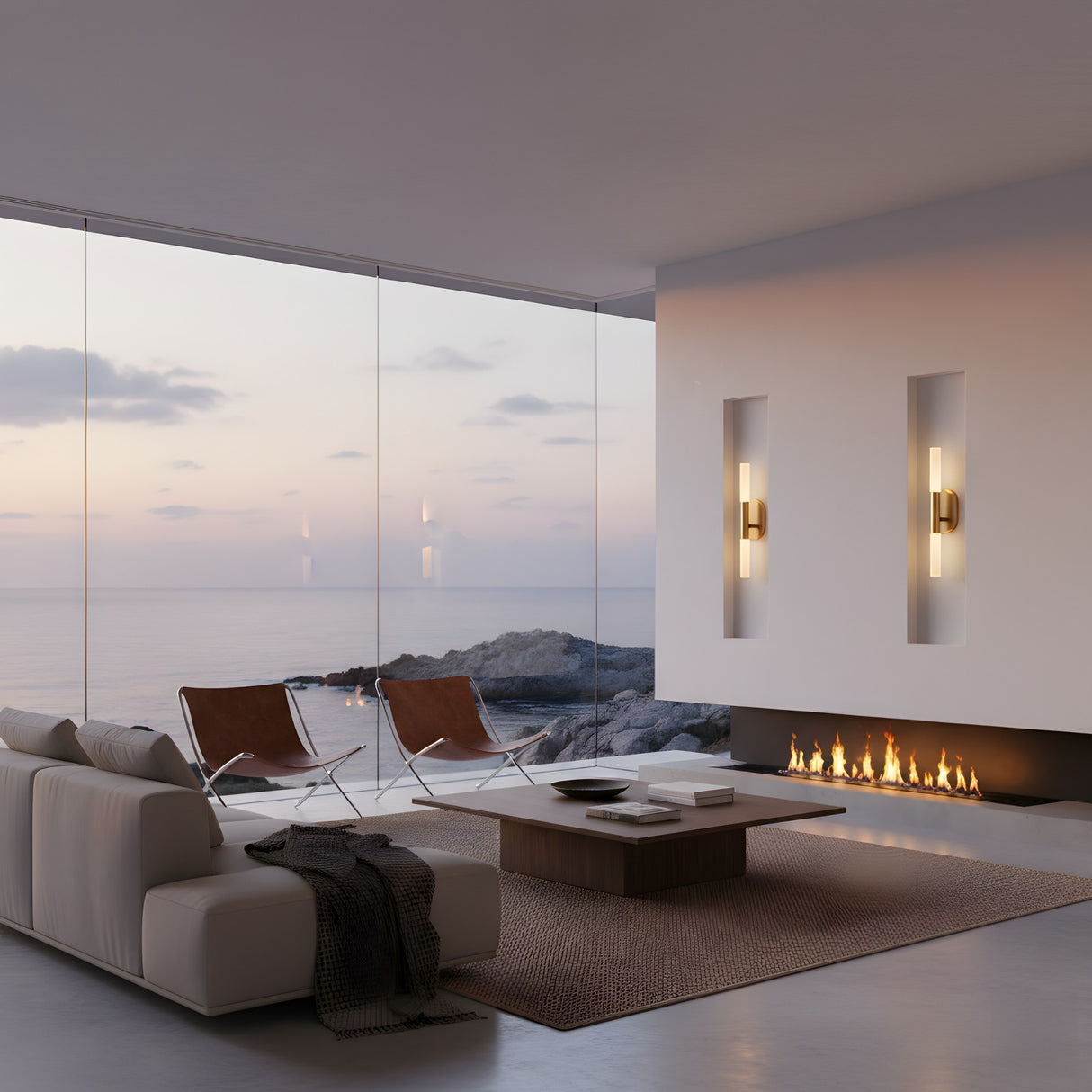A modern living room with floor-to-ceiling windows overlooks a rocky coast at sunset, featuring a white sofa, leather chairs, and a sleek fireplace flanked by Aurelian LED Double Column Alabaster Wall Lights in Brushed Brass & Soft White.