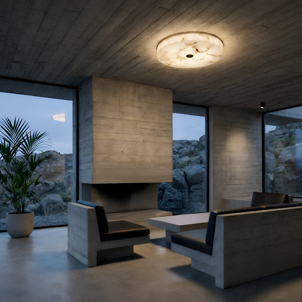 A modern, minimalist living room with concrete walls and ceiling, built-in fireplace, black and gray bench seats around a white table, large windows with rocky views, and a Veltrix LED Small Round Alabaster Flush Ceiling Light in soft white & brushed brass.