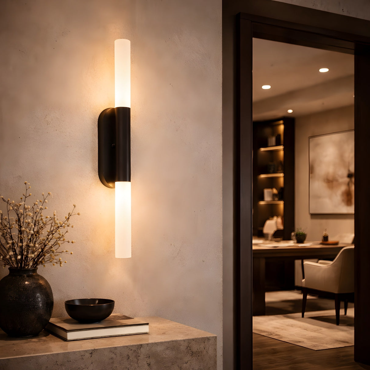 The Aurelian LED Double Column Alabaster Wall Light in Matte Black & Soft White illuminates a neutral wall, accompanied by a stone console with decor; a chic office with art and shelves forms the backdrop.