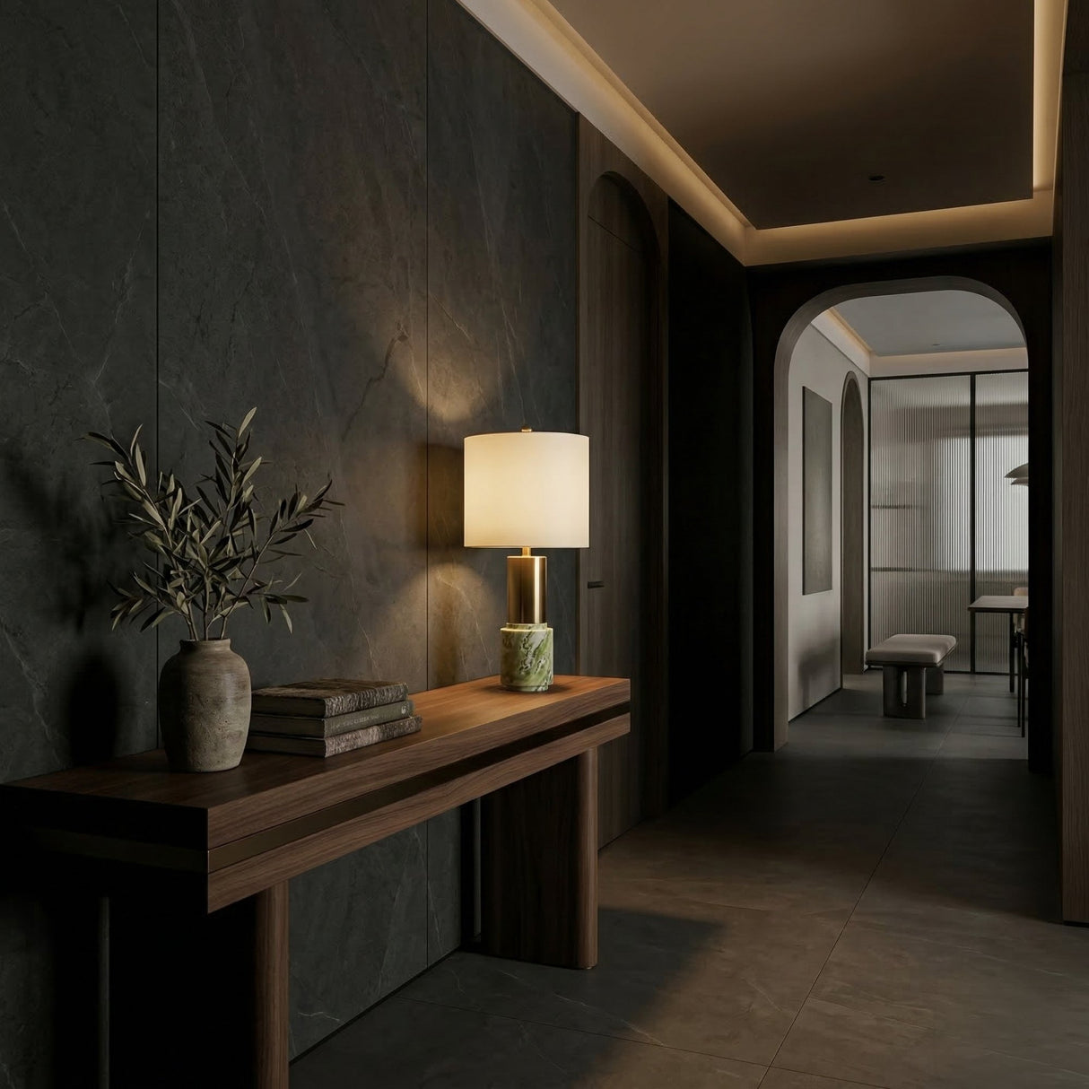 A modern hallway with dark stone walls features a wooden console topped with the Lunaro 1 Light Marble Table Lamp in Brass & Green, books, and a vase with olive branches. Soft lighting and arched doorways lead to a bright room beyond.