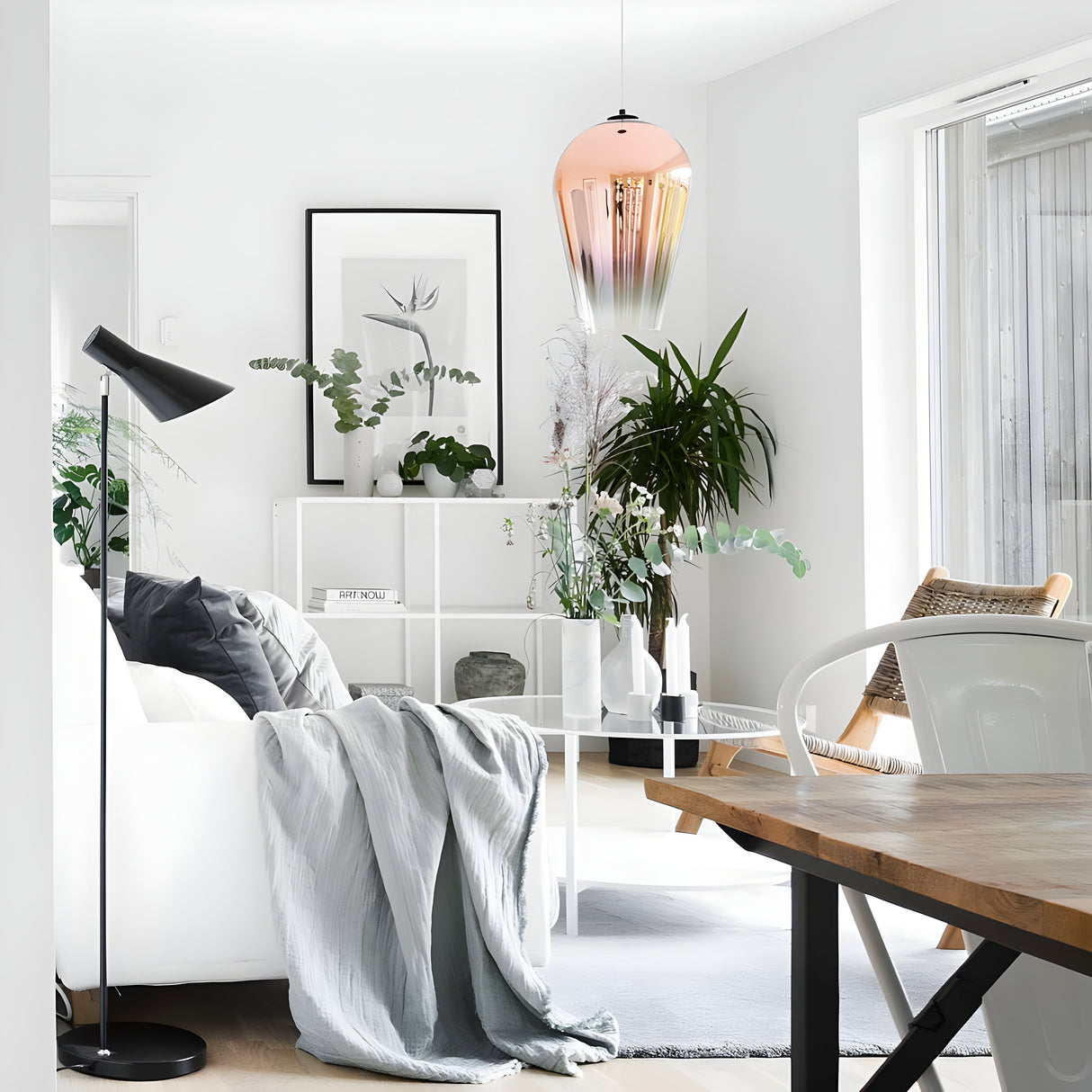 Bright, minimalist living room featuring a white sofa, green plants, and framed artwork. The Slice Glass Pendant Light in a copper finish is elegantly suspended above a wooden dining table. Light floods in from a large window, highlighting the neutral-colored decor.