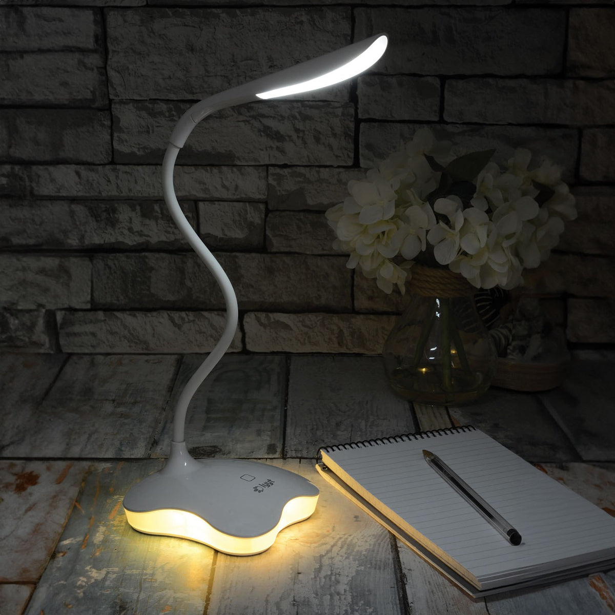 LYYT LED touch sensor USB desk lamp in white with flexible gooseneck illuminated on a wooden surface beside a notepad, pen and vase of white flowers.