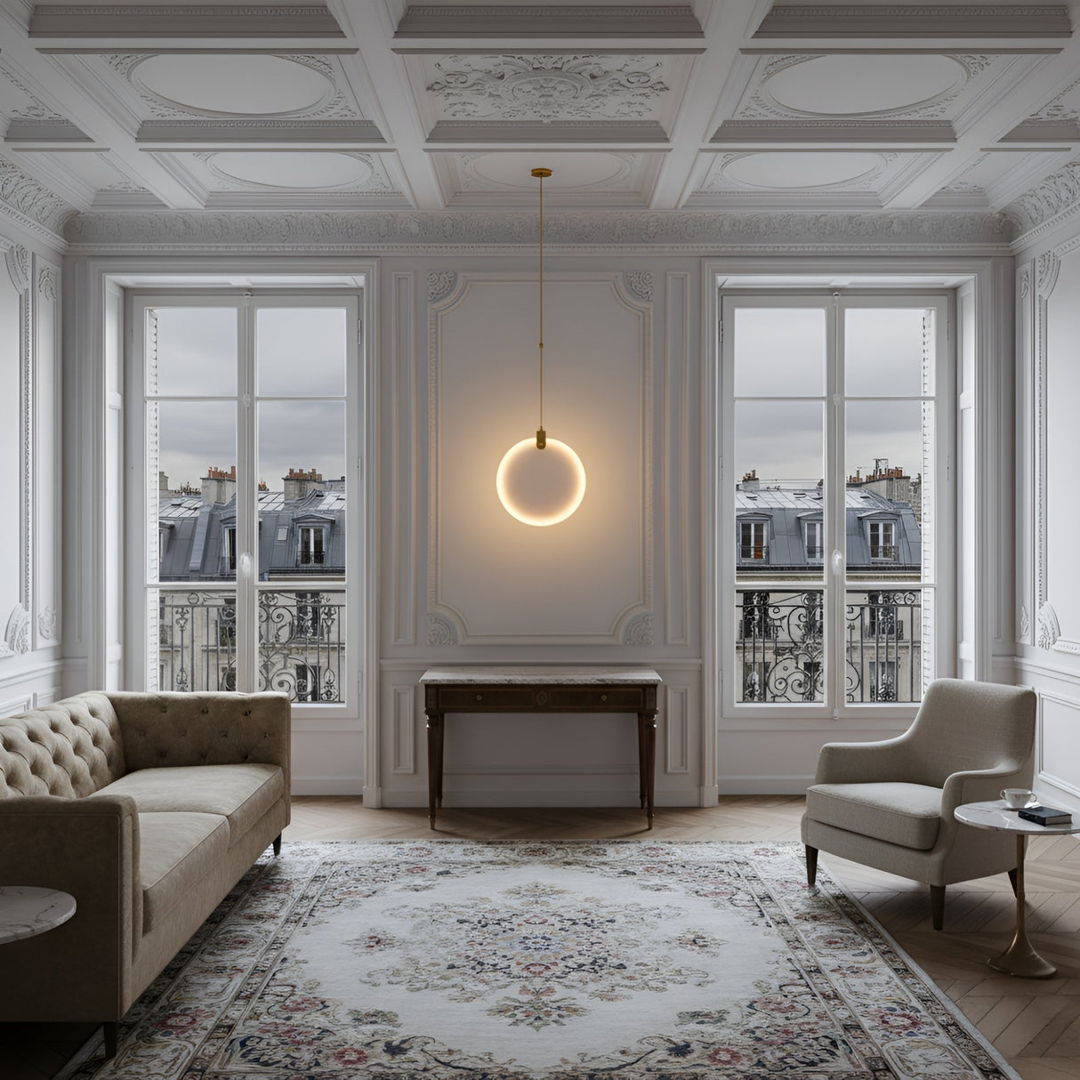 Elegant living room with ornate white walls and ceiling, large windows with Parisian balcony views, a beige sofa, gray armchair, patterned rug, wooden console table, and a Luna LED Medium Alabaster Single Pendant Light - Soft White & Brushed Brass as a centerpiece.