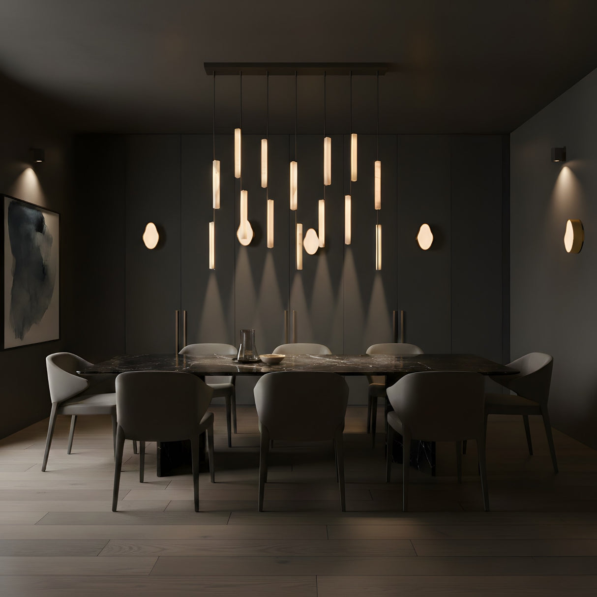 A modern dining room with a dark palette features a long black table, eight grey chairs, abstract wall art, and Marea LED Alabaster Wall Lights in brushed brass above the table, creating a warm, elegant ambiance.