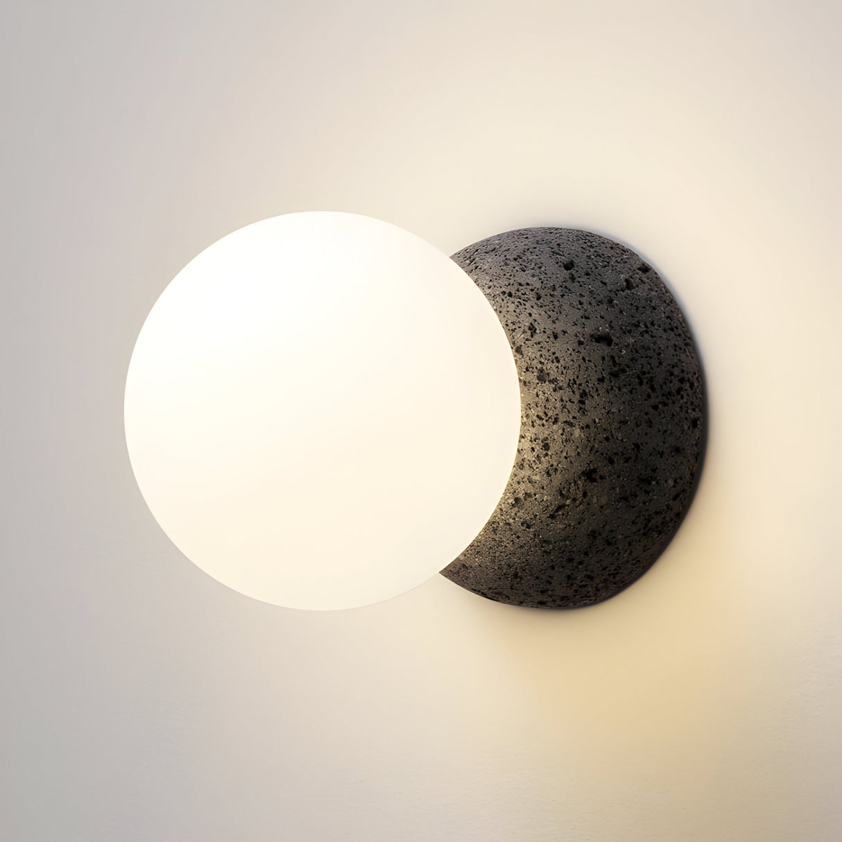 The Origo Concrete Wall Light - Black & White features an opal glass globe diffuser set atop a textured volcanic rock sphere, casting a warm, soft glow against neutral walls.