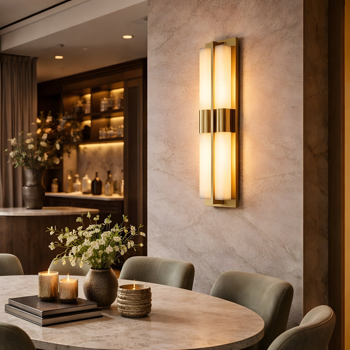 A modern dining room features a round marble table, green chairs, candles, and a floral centerpiece. The Caldera LED Small Split-Core Alabaster Wall Light in brushed brass emits warm light near shelves with decor and bottles.