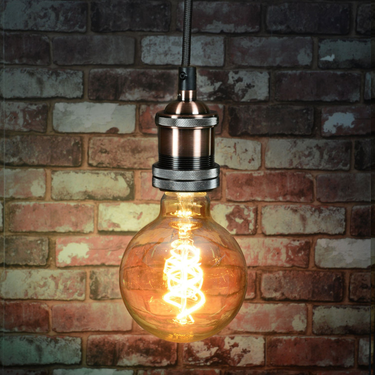 LYYT ES pendant set in antique copper with knurled E27 lamp holder and braided cable, lit amber globe filament bulb against an exposed brick wall.
