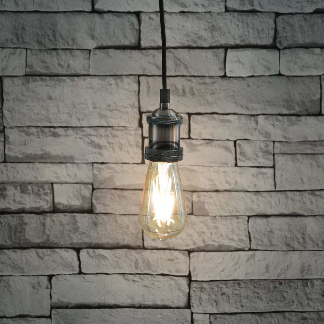 LYYT ES pendant set by AVSL in antique copper, lit E27 filament bulb hanging from braided cable against a white-washed brick wall.